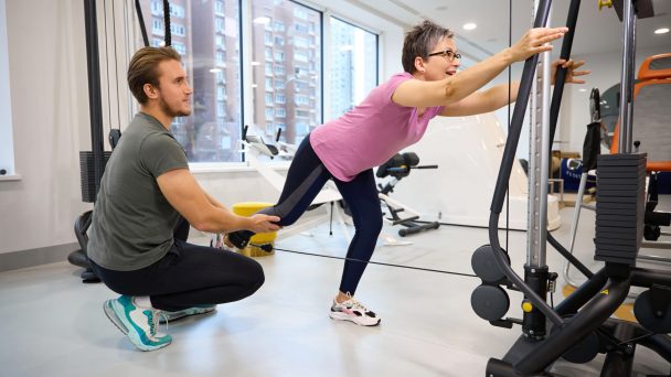 Male trainer helping adult caucasian woman wearing sportswear doing sport exercise for leg in gym. Concept of modern healthy lifestyle